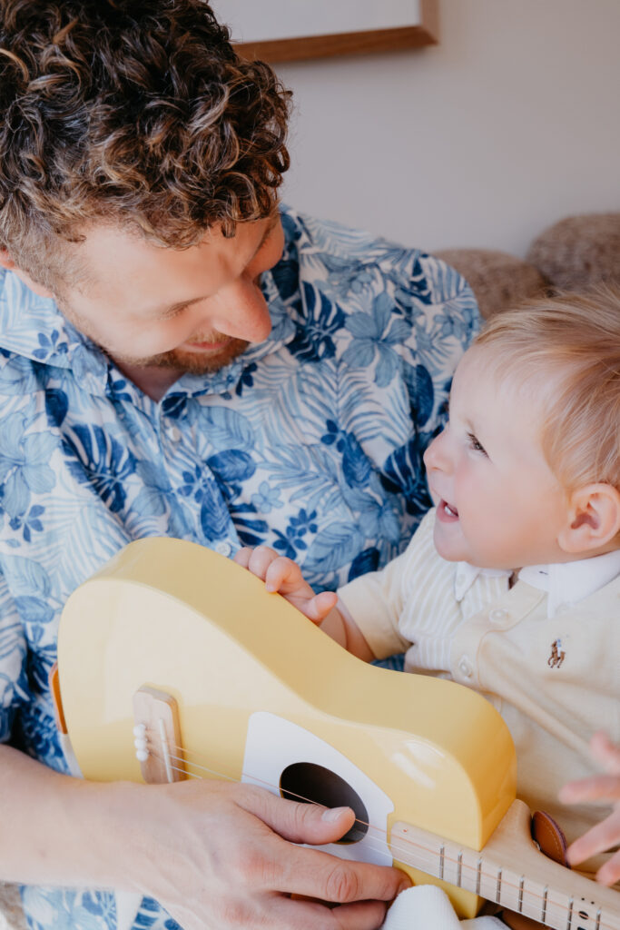 Dad and 9 month old baby son play the yellow guitar together at home during their family photoshoot.