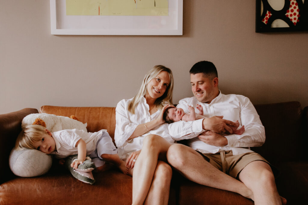Mum, Dad, 2 year old boy and their newborn baby girl sitting on the sofa, wearing tones of white and pink. 