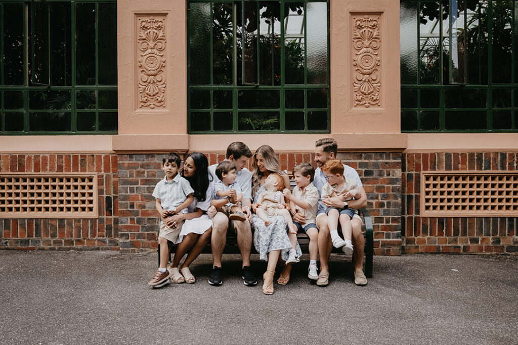 An extended family of four adults and five children sit together on a bench outside the Fitzroy gardens conservatory.